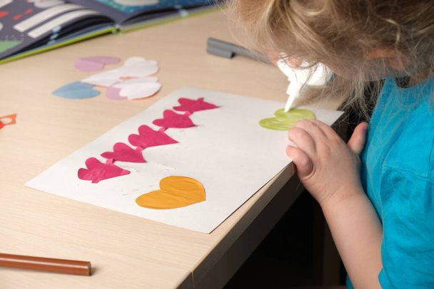 preschooler girl in blue t-shirt makes applique sitting at the table, hearts cut out of colored bougue for card for Valentine's Day, children's creativity, atopic dermatitis on the hands of a child - 写真・画像