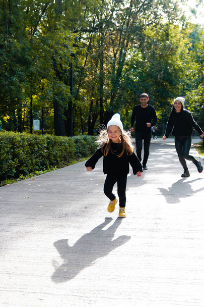 A little girl runs in front, parents, mom and dad run behind. Summer park. Green trees. The concept of a young family.  - 写真・画像