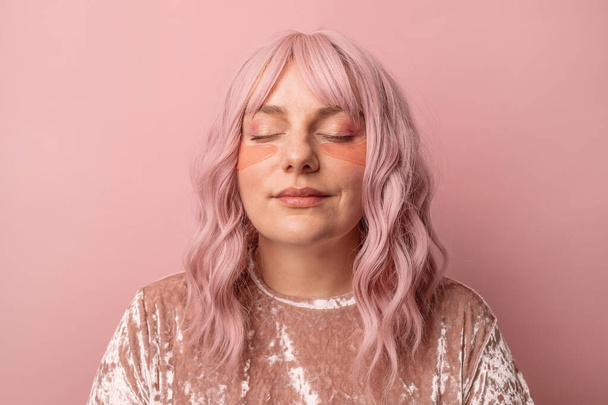 Portrait of happy caucasian female with closed eyes applying pink under eye patches in morning isolated over pink background. Cosmetic care - Foto, Imagen