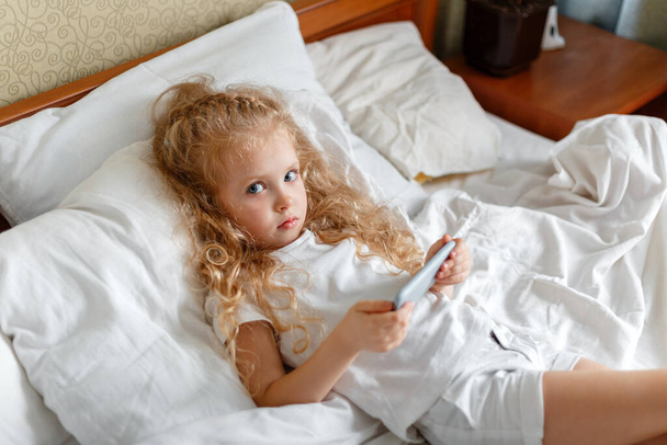 Beautiful Child girl Portrait using smartphone lying in bed. Little blond curly girl play game on smartphone in bedroom at home. Serious offended disgruntled Caucasian Girl use mobile phone - Fotografie, Obrázek