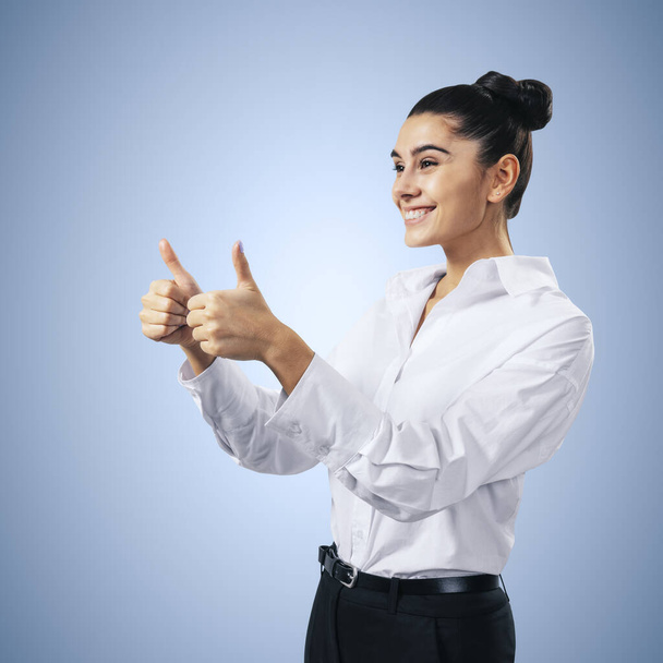Like concept with happy young woman in white shirt holding thumbs up on abstract light blue background - Photo, image