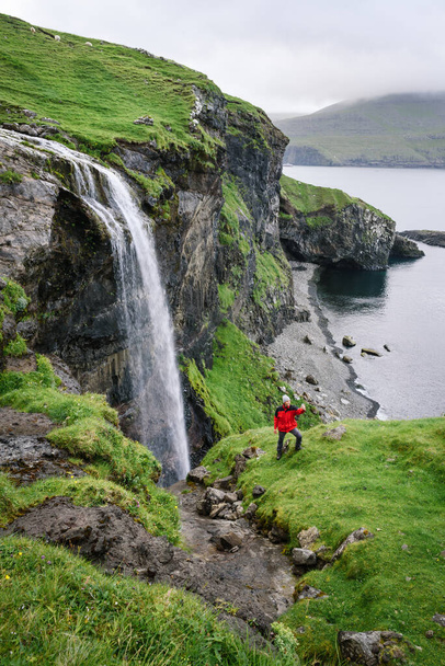 A traveler in a red jacket takes a selfie on the phone near the Skardsafossur waterfall on the island of Vagar, Faroe Islands - Фото, изображение