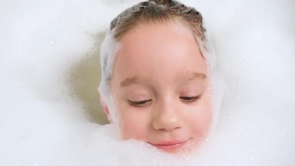 Portrait of cheerful little girl lying in bath with thick soap foam and looking in camera. Concept of child hygiene and health care at home. Child having fun and playing at home - Footage, Video