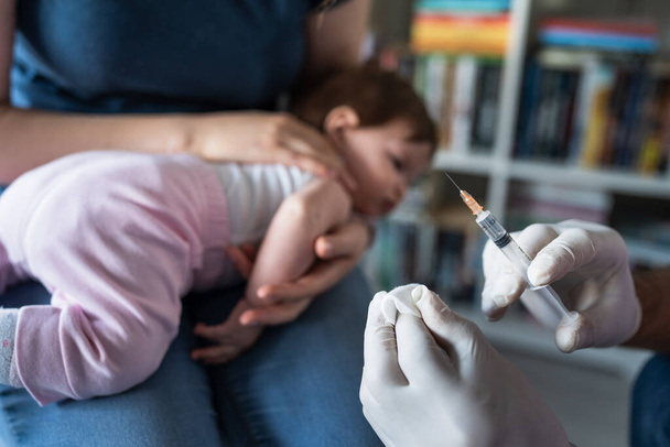 Close up on hand of unknown man doctor wearing protective medical gloves and holding a syringe preparing injection shot with vaccine for small caucasian baby mother hold her child for vaccination - Photo, Image