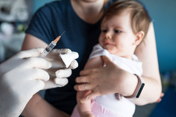 Close up on hand of unknown man doctor wearing protective medical gloves and holding a syringe preparing injection shot with vaccine for small caucasian baby mother hold her child for vaccination - Foto, Imagem