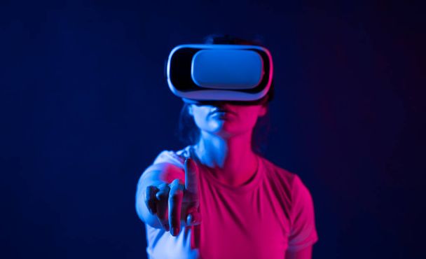 Virtual reality technology concept. Portrait of young woman stands in studio wearing VR glasses and swiping scrolling with hand in air. - 写真・画像