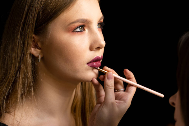 Makeup artist applies red lipstick on a beautiful woman face. Hand of make-up master, painting lips of young beauty model girl. Make up in process. - 写真・画像