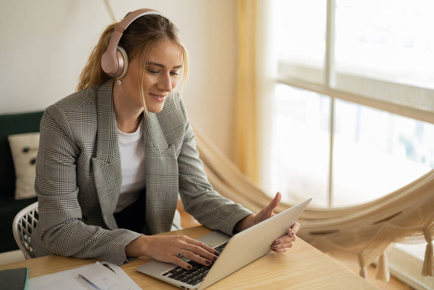 Egy fiatal nő, aki otthon dolgozik laptoppal, papírokkal és fejhallgatóval. Otthoni iroda koncepció. Szürke jegyzetfüzet a munkához. Kiváló minőségű fénykép - Fotó, kép