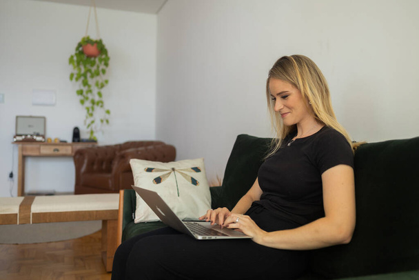 Mujer joven que trabaja en casa desde su sofá de la sala de estar con el ordenador portátil en su regazo. Concepto de oficina en casa. Cuaderno gris para trabajar. Concepto de oficina en casa. Foto de alta calidad - Foto, Imagen