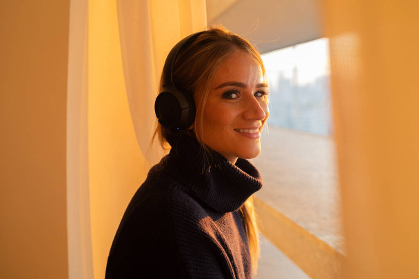 Side view of young woman listening to music with headphones in her ears by the window with a city view landscape in the background. High quality photo. Mobile phone, technology, music, urban living, feel good concept. - Foto, Bild