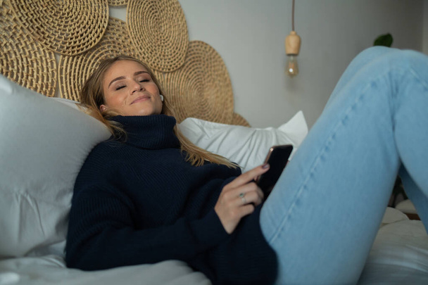 Mladá žena poslouchá hudbu se sluchátky v uších v posteli v ložnici ve svém bytě. Kvalitní fotka. Mobilní telefon, technologie, hudba, městský život, dobrý koncept. - Fotografie, Obrázek