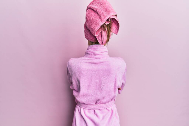 Mujer rubia joven con gorra de toalla de ducha y albornoz mirando hacia atrás con los brazos cruzados  - Foto, Imagen