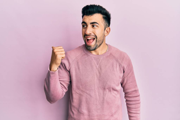 Νεαρός Ισπανός που φοράει καθημερινά ρούχα δείχνοντας τον αντίχειρα στο πλάι χαμογελώντας χαρούμενος με ανοιχτό στόμα  - Φωτογραφία, εικόνα