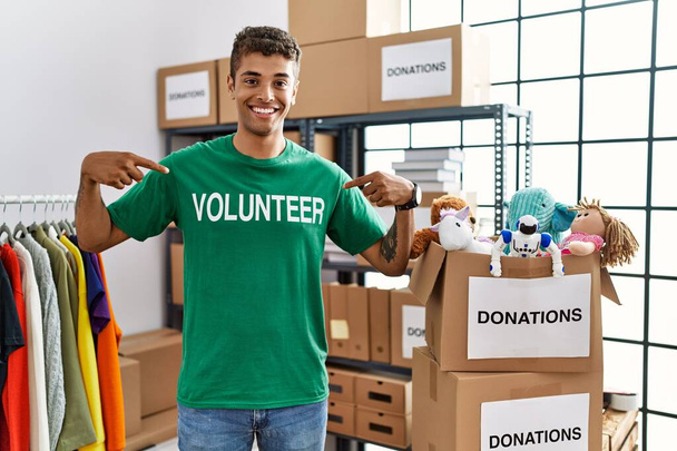 Junger gutaussehender hispanischer Mann im freiwilligen T-Shirt am Spendenstand sieht selbstbewusst aus mit einem Lächeln auf dem Gesicht, zeigt mit den Fingern stolz und glücklich.  - Foto, Bild