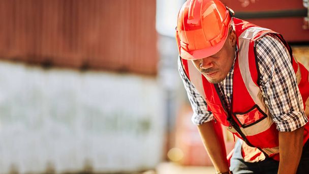 Afro-Amerikaanse arbeiders zijn uitgeput door hard werken. Een container werf werknemer draagt een rode reflecterende vest en harde hoed staat te moe als gevolg van het warme weer. - Foto, afbeelding