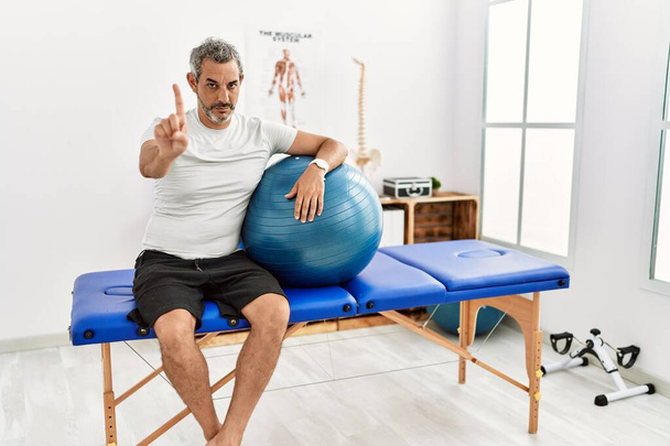 Middle age hispanic man at pain recovery clinic holding pilates ball pointing with finger up and angry expression, showing no gesture  - Photo, Image