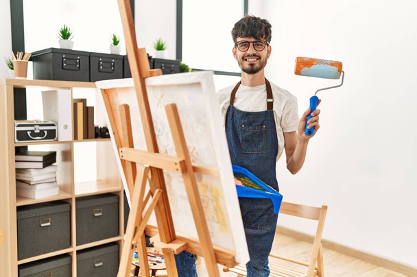 Nuori latino taiteilija mies hymyilee onnellinen piirustus käyttäen maalaustela on art studio. - Valokuva, kuva