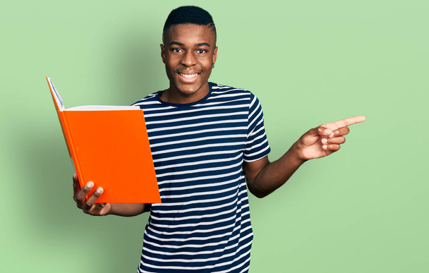 Jeune homme afro-américain tenant livre souriant heureux pointant avec la main et le doigt sur le côté  - Photo, image