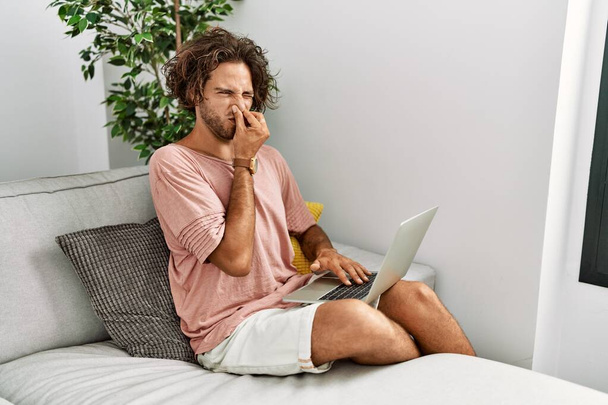 Молодий іспаномовний чоловік сидить на дивані вдома, використовуючи ноутбук пахне чимось огидним і огидним, нестерпним запахом, тримаючи дихання пальцями на носі. поганий запах
  - Фото, зображення
