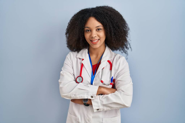 Jonge Afro-Amerikaanse vrouw in doktersuniform en stethoscoop blij gezicht glimlachend met gekruiste armen kijkend naar de camera. positieve persoon.  - Foto, afbeelding