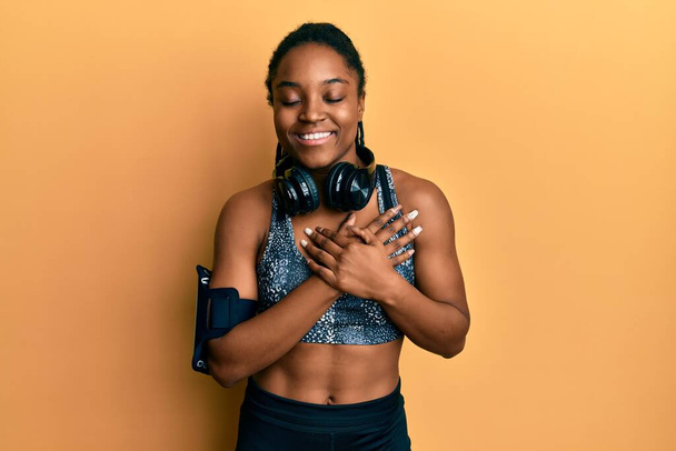 African american woman with braided hair wearing sportswear and arm band smiling with hands on chest with closed eyes and grateful gesture on face. health concept.  - Foto, afbeelding