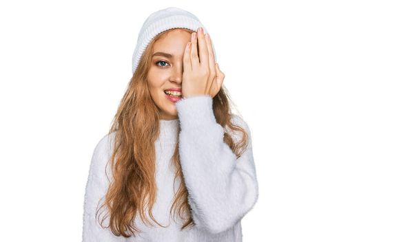 Молода біла дівчина у вовняному светрі та зимовій кепці, що покриває одне око рукою, впевнена посмішка на обличчі та несподівані емоції
.  - Фото, зображення