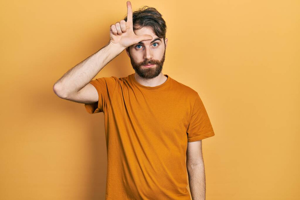 Hombre caucásico con barba usando camiseta amarilla casual burlándose de la gente con los dedos en la frente haciendo gesto perdedor burlándose e insultando.  - Foto, Imagen