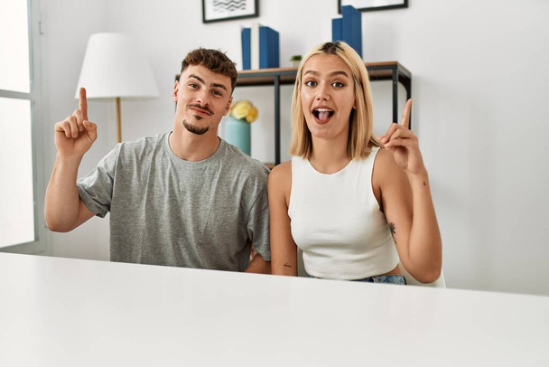 Ein junges schönes Paar in lässiger Kleidung sitzt zu Hause auf dem Tisch und zeigt mit erhobenem Zeigefinger auf eine erfolgreiche Idee. aufgeregt und glücklich. Nummer eins.  - Foto, Bild