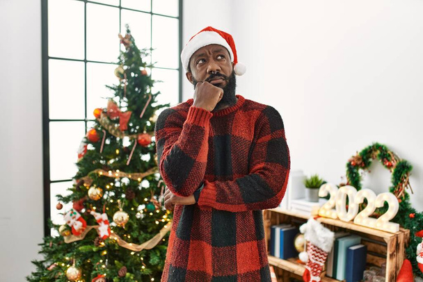 Afrikai amerikai férfi télapó sapkát visel, karácsonyfa mellett áll, kezével az állán, gondolkodva a kérdésen, töprengő kifejezésen. elmosolyodott, figyelmes arccal. kétség.  - Fotó, kép