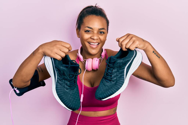 Young african american girl wearing gym clothes, using headphones and holding sporty shoes smiling and laughing hard out loud because funny crazy joke.  - Foto, immagini