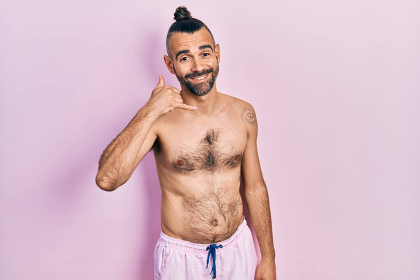Fiatal spanyol férfi félmeztelenül fürdőruhát visel, mosolyog, telefonos mozdulatokat tesz, kezével és ujjaival, mintha telefonálna. kommunikációs koncepciók.  - Fotó, kép