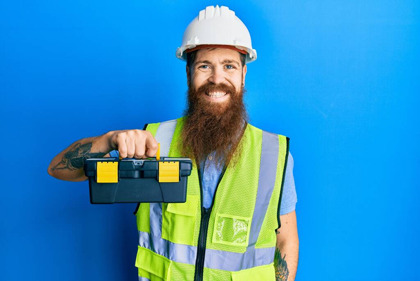 Hombre pelirrojo con barba larga con casco de seguridad y chaqueta reflectante sosteniendo caja de herramientas que se ve positivo y feliz de pie y sonriendo con una sonrisa confiada mostrando los dientes  - Foto, imagen