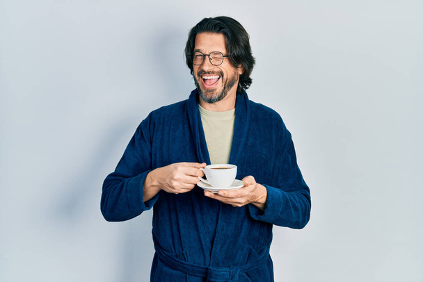 Middle age caucasian man wearing robe drinking coffee winking looking at the camera with sexy expression, cheerful and happy face.  - Foto, immagini