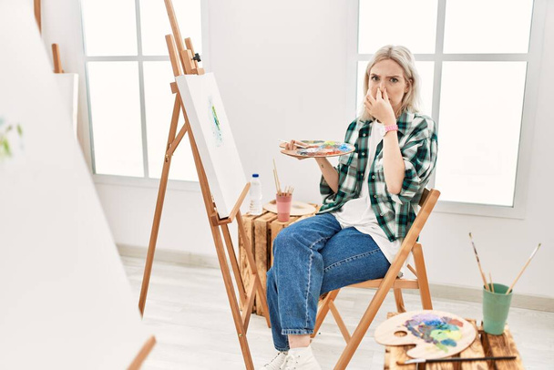 Young artist woman painting on canvas at art studio smelling something stinky and disgusting, intolerable smell, holding breath with fingers on nose. bad smell  - Foto, Imagem