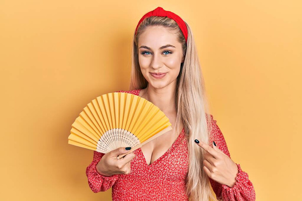 Jeune femme caucasienne agitant ventilateur main refroidissement de l'air en été souriant heureux pointant avec la main et le doigt  - Photo, image