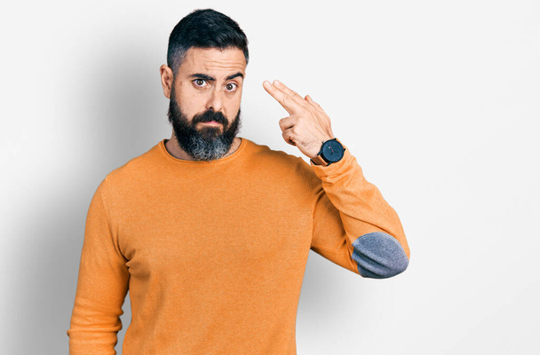 Hombre hispano con barba vistiendo suéter casual de invierno disparando y matándose apuntando de la mano y los dedos a la cabeza como arma, gesto suicida.  - Foto, imagen