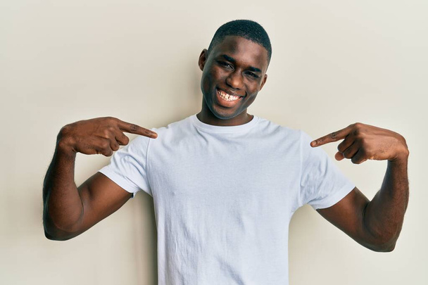Genç Afro-Amerikan adam, günlük beyaz tişört giyiyor. Kendine güvenen, yüzünde gülümseme olan, kendini gururlu ve mutlu bir şekilde gösteren..  - Fotoğraf, Görsel