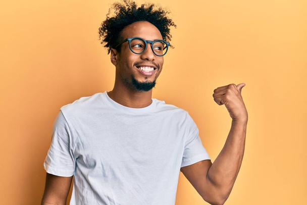 Jovem afro-americano com barba vestindo camisa branca casual e óculos apontando o polegar para o lado sorrindo feliz com a boca aberta  - Foto, Imagem
