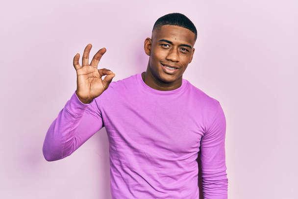 Молодой черный мужчина в обычном розовом свитере с положительной улыбкой делает знак ОК с рукой и пальцами. успешное выражение.  - Фото, изображение