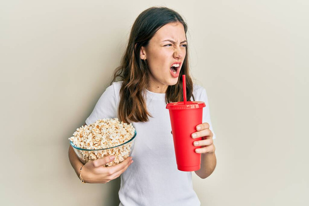 Giovane donna bruna mangiare popcorn e bere soda arrabbiato e pazzo urlando frustrato e furioso, gridando con rabbia. rabbia e concetto aggressivo.  - Foto, immagini