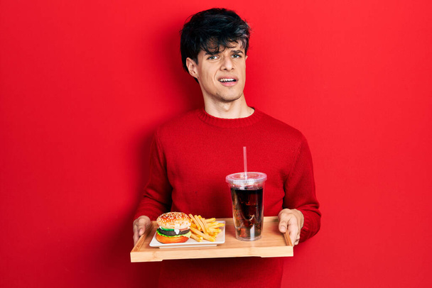 Jóképű hipszter fiatalember eszik egy ízletes klasszikus hamburgert sült krumplival és szódával, tudatlan és zavaros kifejezéssel. kétség.  - Fotó, kép