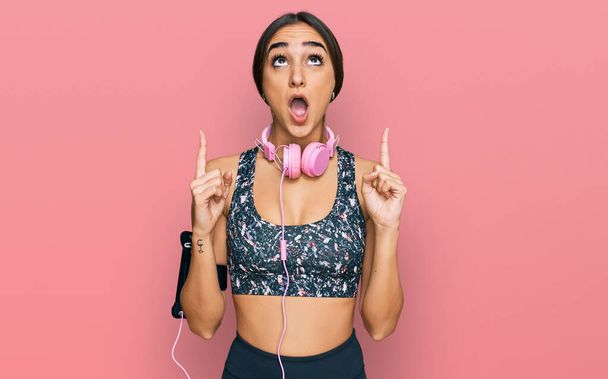 Beautiful brunette woman wearing gym clothes and using headphones amazed and surprised looking up and pointing with fingers and raised arms.  - Photo, Image