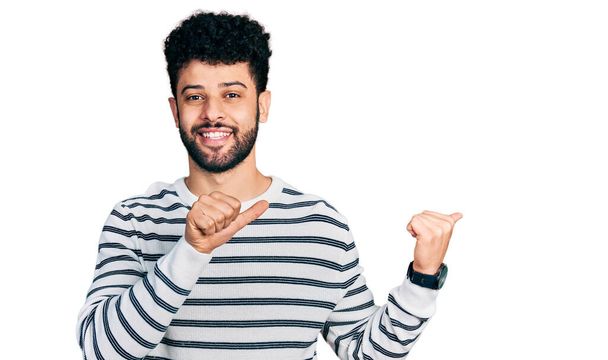 自信を笑顔、手と親指で後ろを指しているカジュアルなストライプのセーターを身に着けている髭を生やして若いアラブ人男性  - 写真・画像