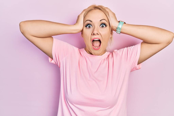 Mladá blondýna nosí ležérní růžové tričko šílené a strach s rukama na hlavě, strach a překvapení z šoku s otevřenými ústy  - Fotografie, Obrázek