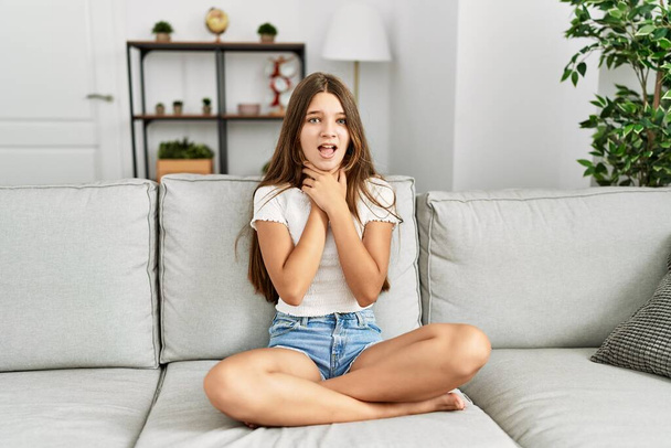 Young brunette teenager sitting on the sofa at home shouting and suffocate because painful strangle. health problem. asphyxiate and suicide concept.  - 写真・画像