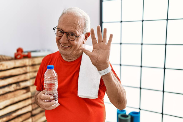 Hombre mayor con ropa deportiva y toalla en el gimnasio renunciando a decir hola feliz y sonriente, gesto de bienvenida amistoso  - Foto, Imagen