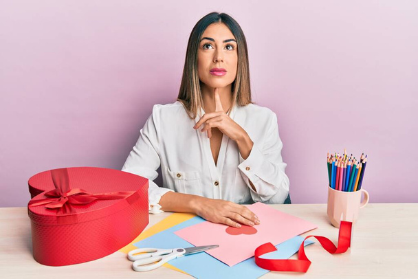 Junge hispanische Frau, die Valentinstagsgeschenk macht, sitzt auf dem Tisch und denkt konzentriert mit dem Finger am Kinn über Zweifel nach und blickt verwundert auf  - Foto, Bild