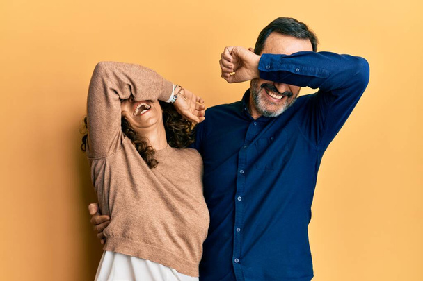 Ein hispanisches Paar mittleren Alters in lässiger Kleidung lächelt fröhlich spielend und guckt ein Buh mit Händen, die Gesicht zeigen. überrascht und aufgeregt  - Foto, Bild