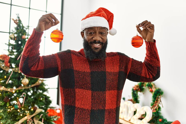 Young african american man smiling confident holding balls standing by christmas hat at home - Photo, Image
