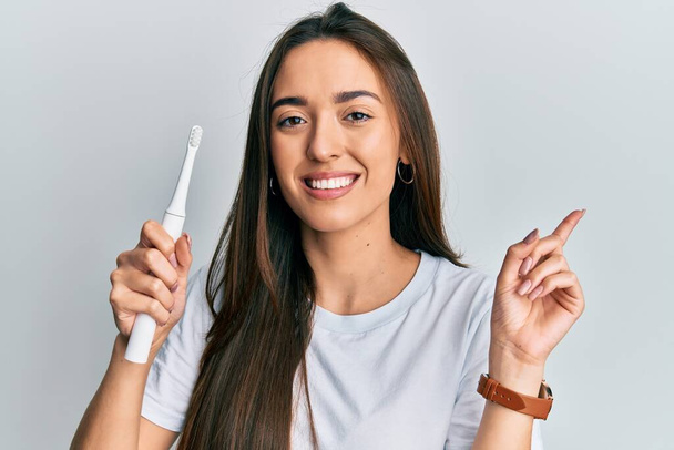 Joven chica hispana sosteniendo cepillo de dientes eléctrico sonriendo feliz señalando con la mano y el dedo hacia un lado  - Foto, Imagen
